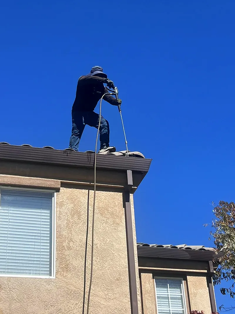 Crew member on a tile roof pressure washing it against a clear blue sky