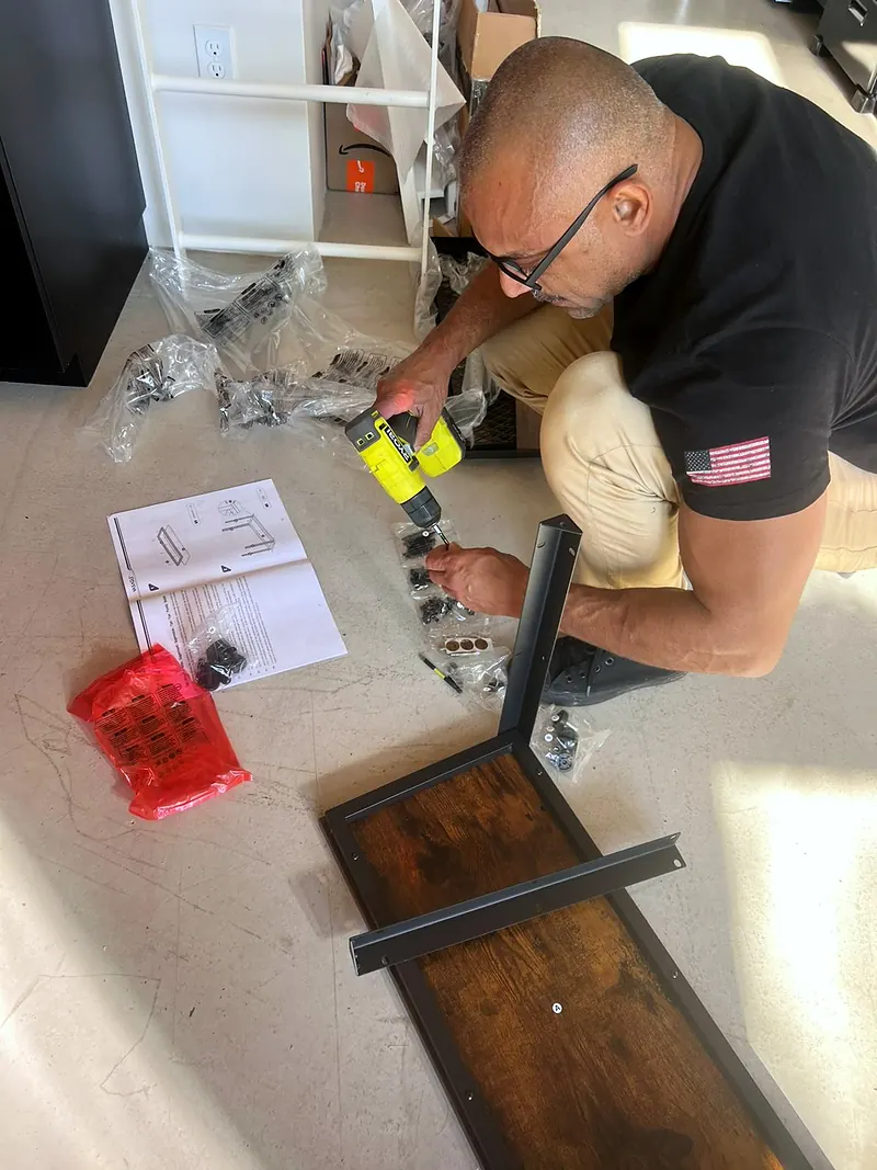 Junior with a cordless drill beside an upside-down wood-top bench, hardware bags and the manual on the floor